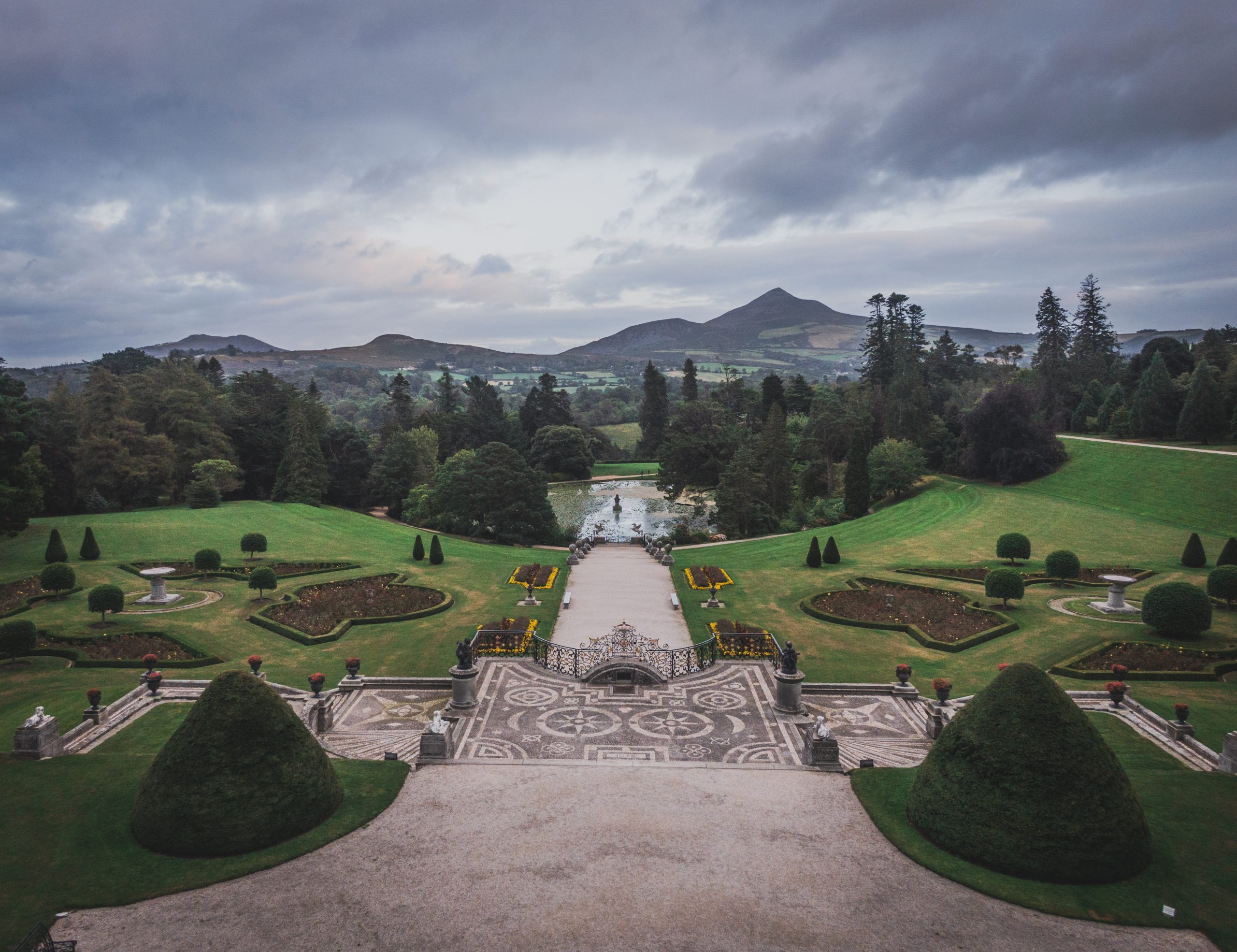 Illustration of a girl in a black cape standing on the Italian Gardens terrace at Powerscourt, looking towards the fountain, Triton Lake and the Sugarloaf Mountain. AI generated